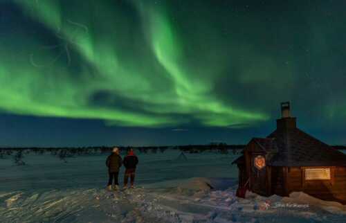 Two people out in the snow next to the small cabin watching the northern lights.