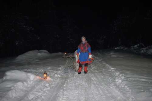 Woman dressed in Sami clothing walking on snowy road with reindeer.