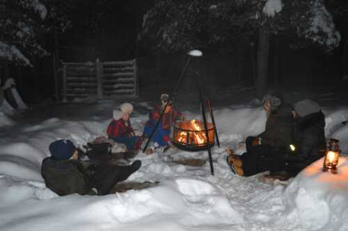 Woman and girl dressed in Sami clothing sitting around a campfire with guests in the snow.
