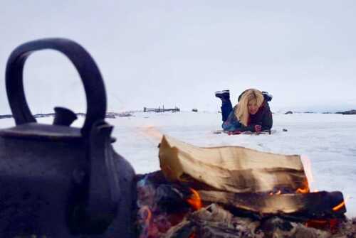 Coffee pot on bonfire in the foreground, woman lying on the snow behind.